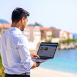 Un agent immobilier consulte un site web immobilier sur son ordinateur portable, en terrasse, avec vue sur la mer et des immeubles typiques du sud de la France en arrière-plan. Soleil, palmiers et ambiance professionnelle.